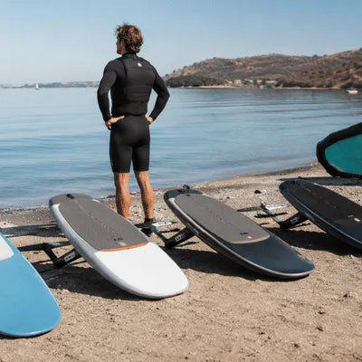 A rider stands on a beach beside several wing foil board options while looking out at calm water