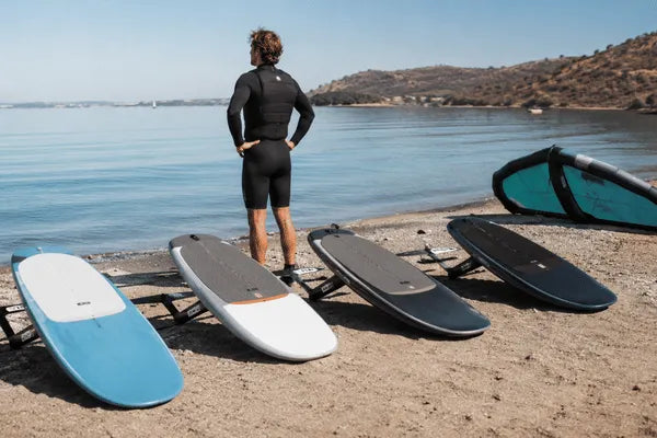 A rider stands on a beach beside several wing foil board options while looking out at calm water