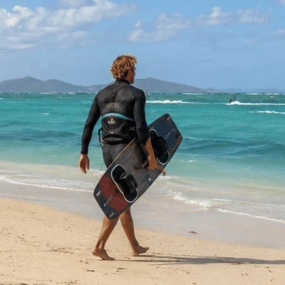 A kiteboarder carries a board along a sandy beach while kiteboarding kites fly over the windy turquoise water
