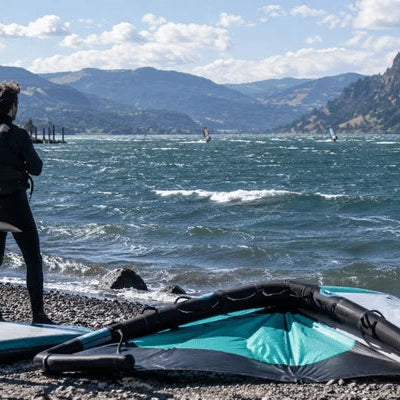 A rider stands beside wing foil gear while checking rough river conditions for a Hood River wind forecast before launching
