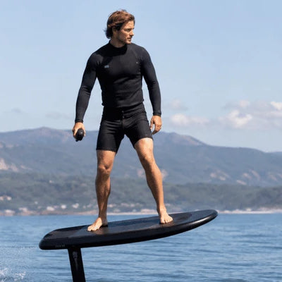 A rider stands balanced on an e foil board lifted above calm water while holding a handheld remote with mountains in the background