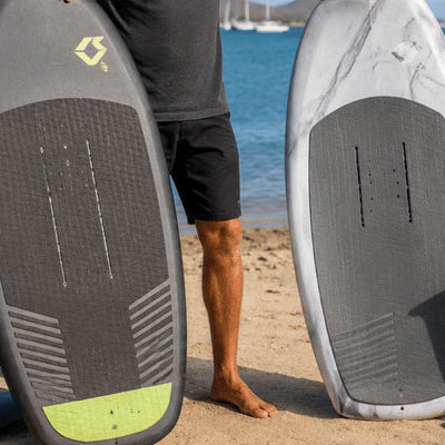 A rider compares two upright cheap foil board options on a sandy beach with foil gear beside the water
