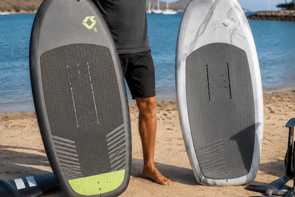 A rider compares two upright cheap foil board options on a sandy beach with foil gear beside the water