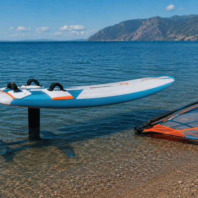 A windsurfing foil board rests in shallow water near the shore with a windsurf sail beside it
