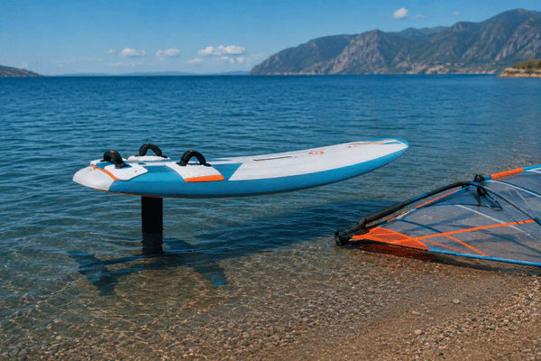 A windsurfing foil board rests in shallow water near the shore with a windsurf sail beside it
