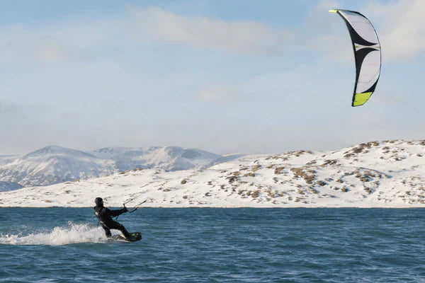 A rider enjoys kite kitesurfing across cold blue water beside snow-covered mountains beneath a large airborne kite