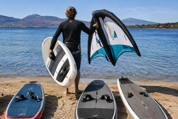 This realistic photo shows a rider choosing a wing foil board from several options on a beach before entering the water